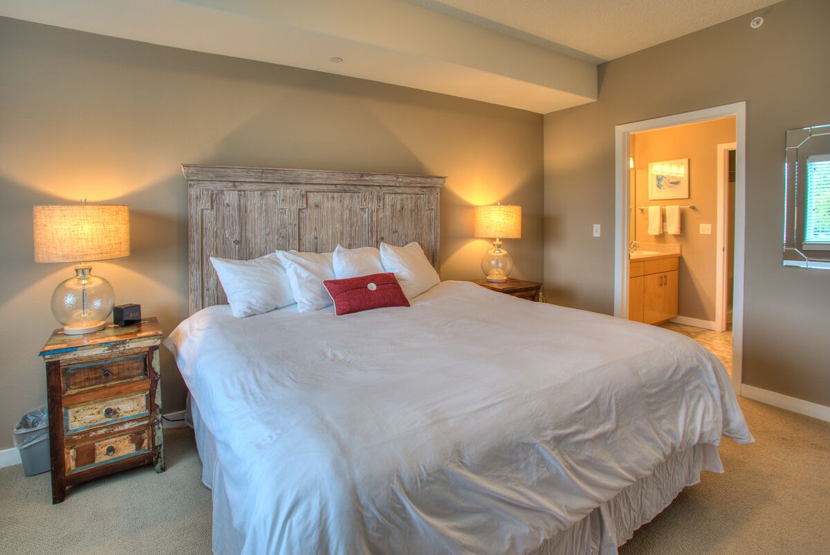 A bedroom with a large bed, two bedside tables with lamps, a wooden headboard, and a door leading to a bathroom.