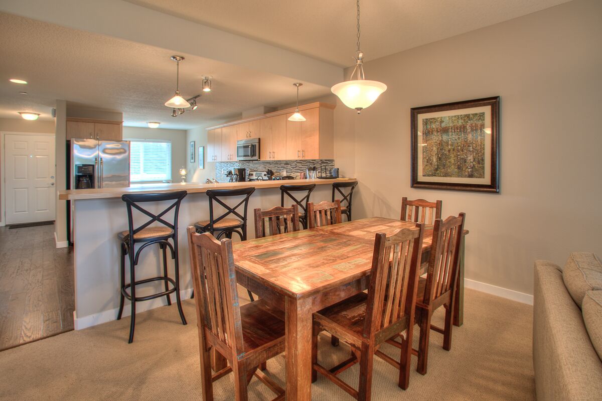 A dining area with a wooden table and chairs, a kitchen in the background, and a framed picture on the wall.