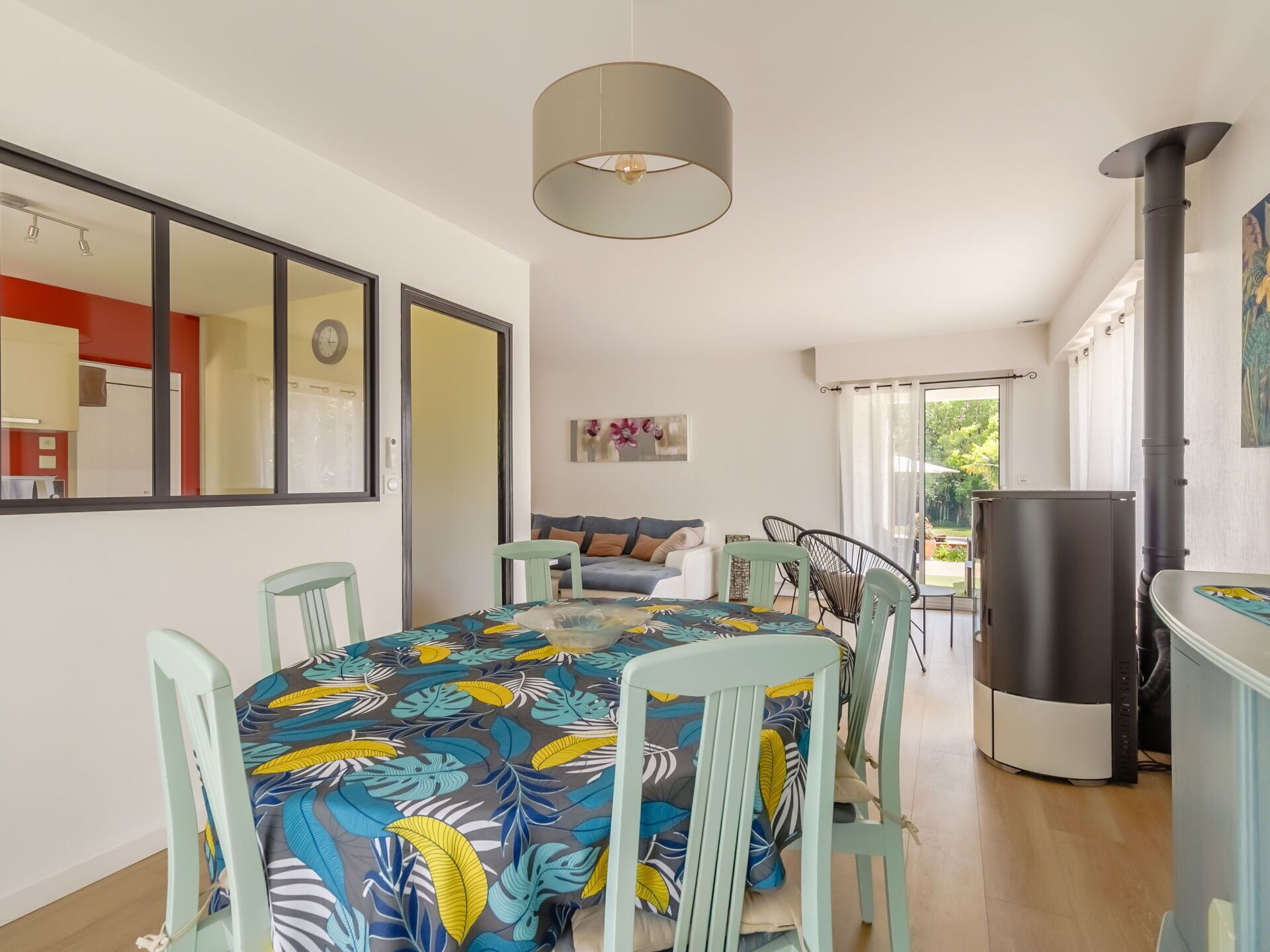 A dining area with a table covered in a colorful leaf-patterned tablecloth, surrounded by light green chairs. A modern pendant light hangs above the table.