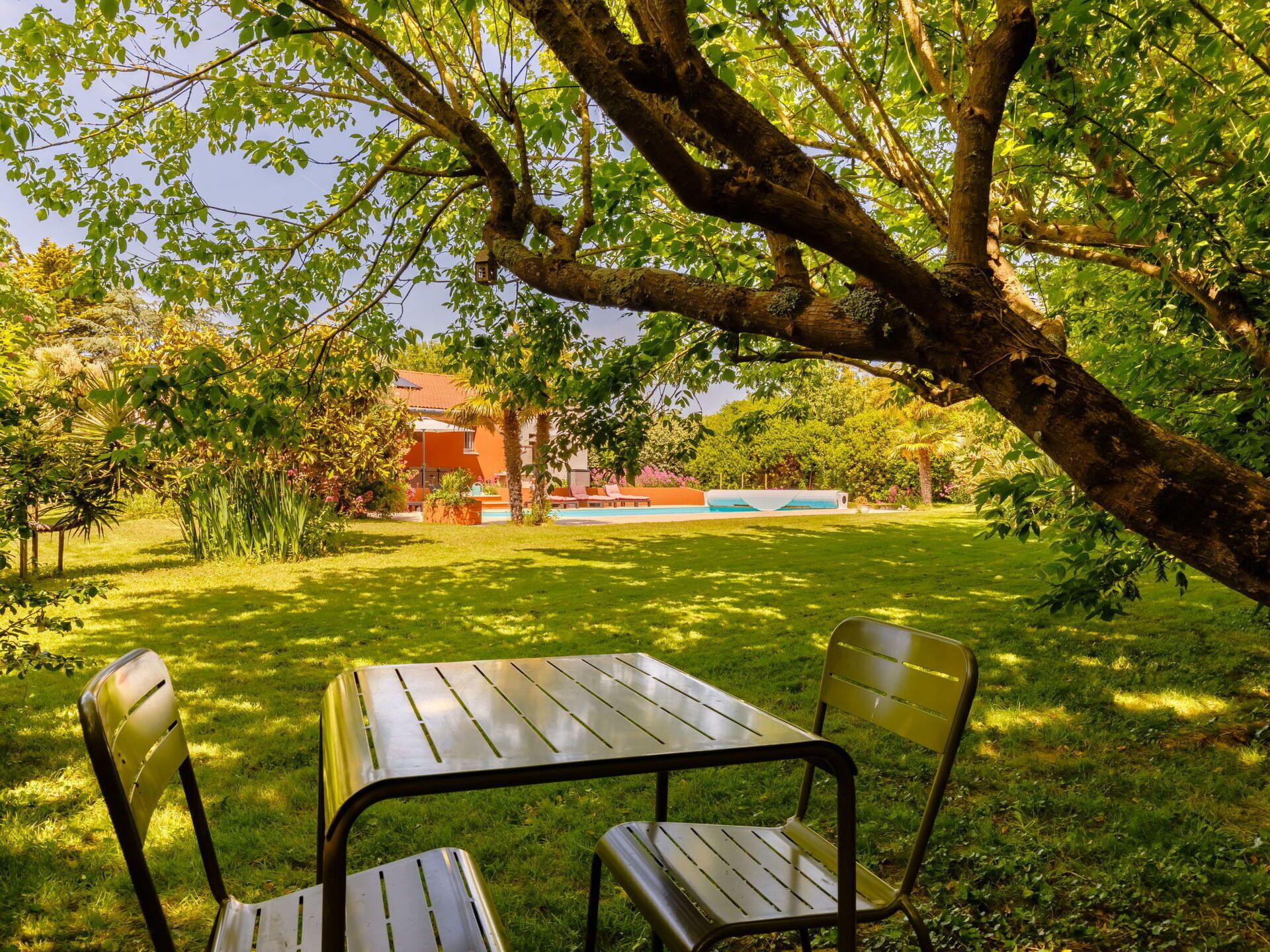 A patio with a table and chairs under a tree, a swimming pool, and a building in the background.