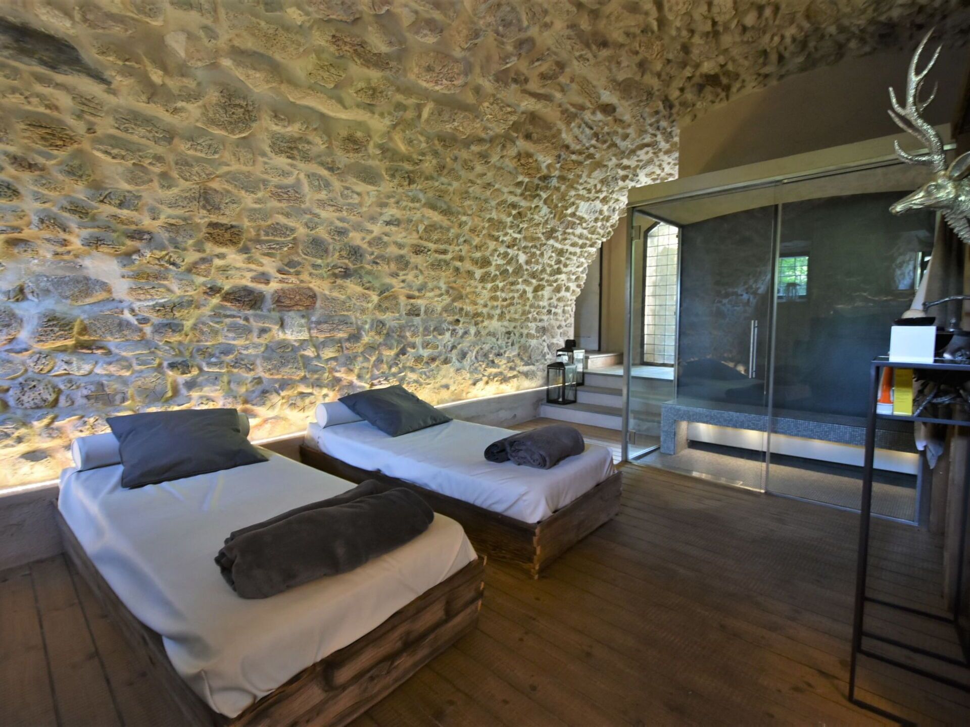 A room with stone walls, two wooden beds with pillows, a glass door leading outside, and a deer head decoration.