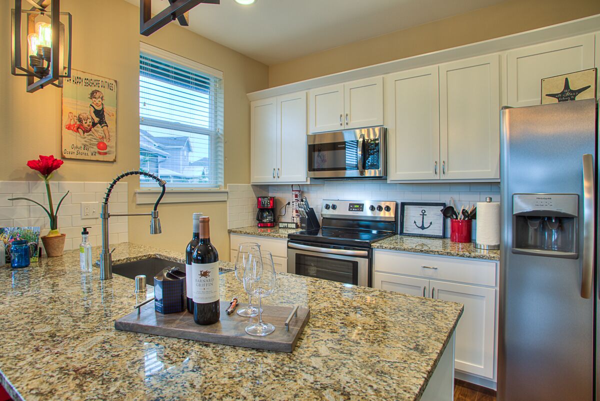 Cocina con armarios blancos, electrodomésticos de acero inoxidable, encimera de granito y una pequeña isla con botellas de vino y copas.