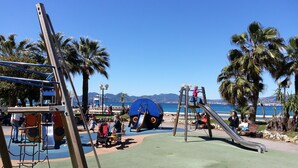 Un terrain de jeux avec des toboggans et des structures d’escalade, entouré de palmiers et offrant une vue dégagée sur la mer.