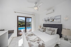 A bedroom with a bed, a nightstand, a lamp, a ceiling fan, a painting, and a view of a swimming pool.