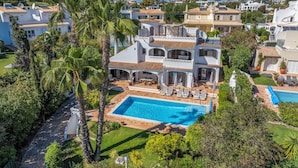 Ein zweistöckiges Haus mit Schwimmbecken, umgeben von üppiger Vegetation und weiteren Häusern.