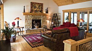 A cozy living room with a stone fireplace, wooden ceiling, and large windows.