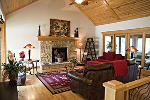 A cozy living room with a stone fireplace, wooden ceiling, and large windows.