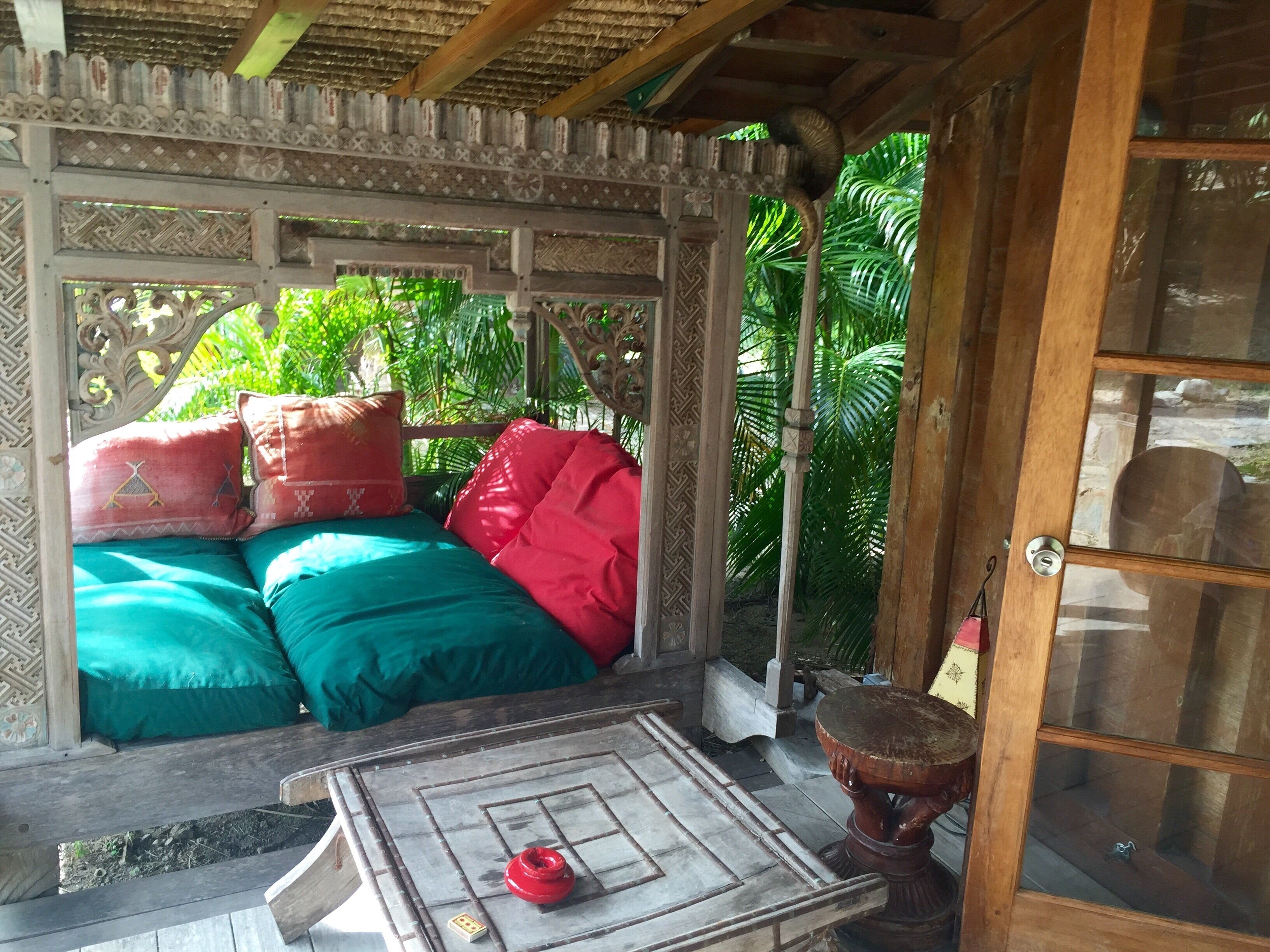 Une pièce traditionnelle en bois, avec un toit de chaume, un lit recouvert d’une literie verte et rouge, et une petite table sur laquelle se trouve un objet rouge.
