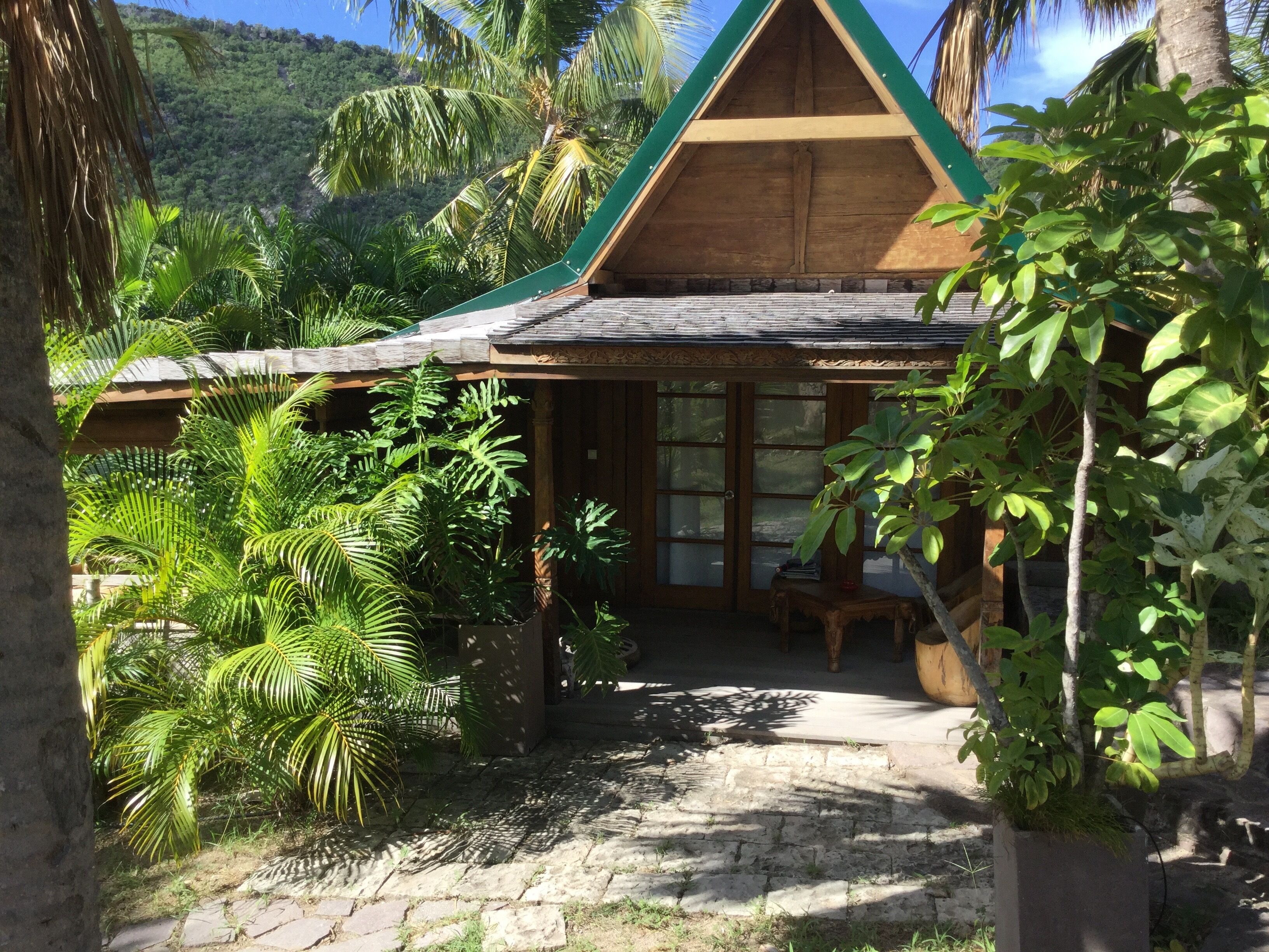 A wooden house with a sloped roof, surrounded by lush greenery and palm trees.