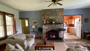 A living room with a stone fireplace, a ceiling fan, and a view into the kitchen.