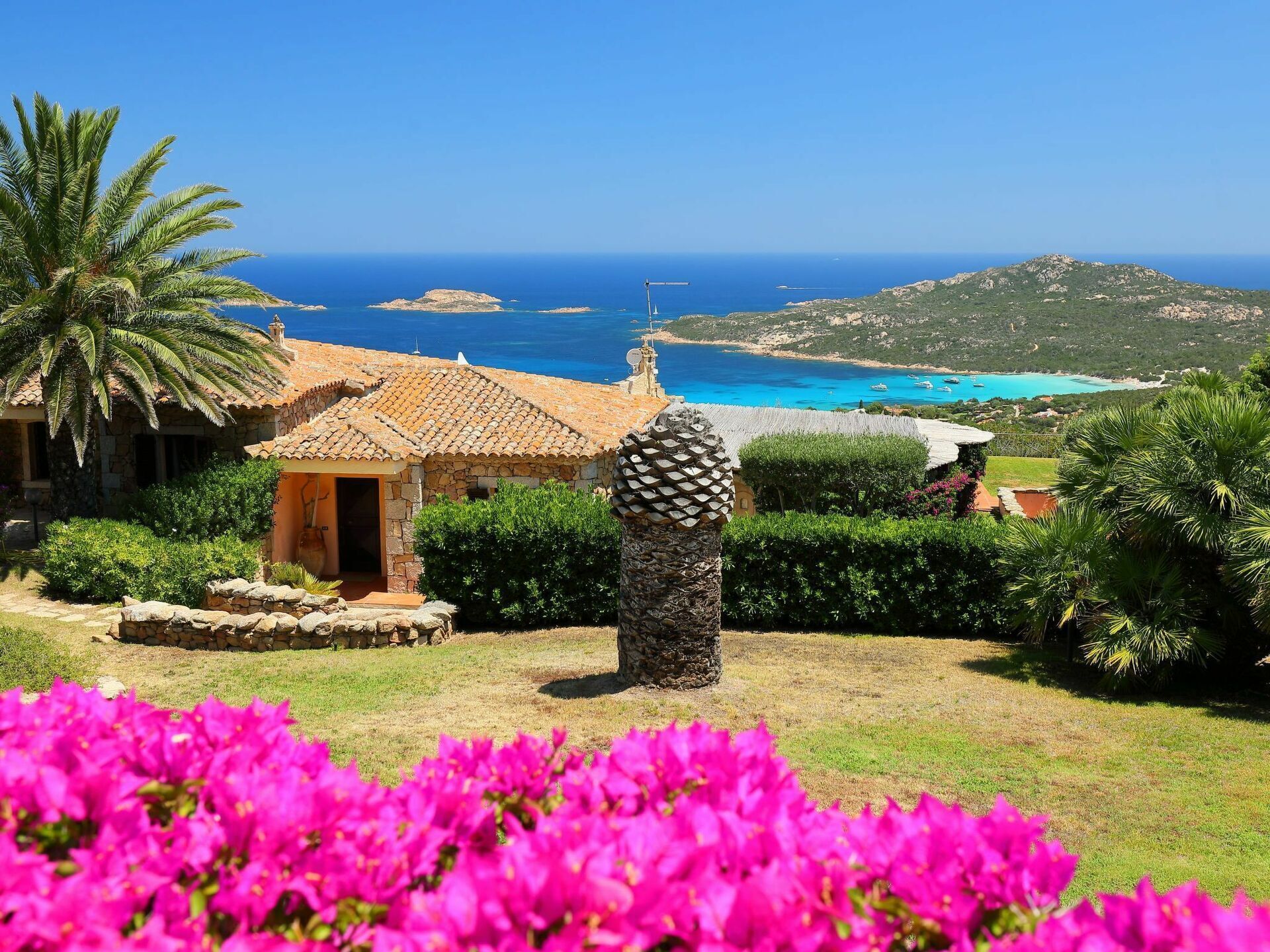 A coastal property with a stone house, a decorative stone pillar, and vibrant pink flowers in the foreground.