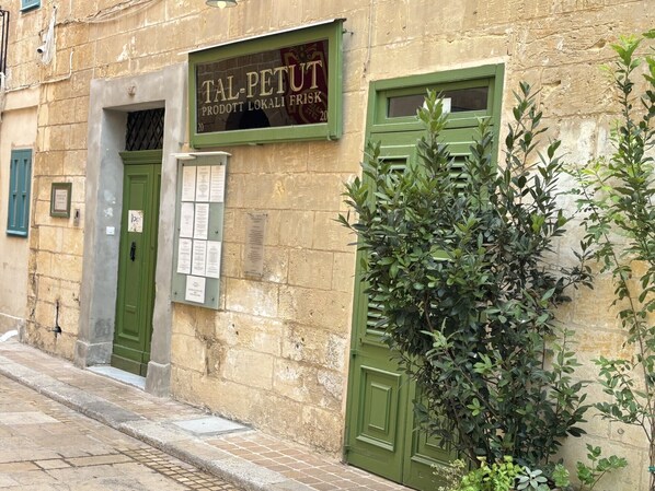 A building with a green door and windows, a sign reading "TAL-PETUT PRODOTT LOKALI FRISK", and a plaque on the wall.