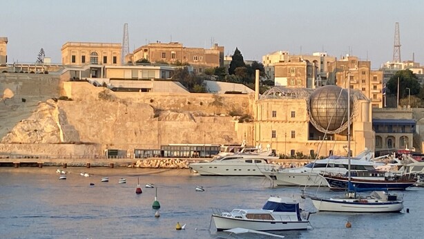 Un port de plaisance avec des bateaux amarrés, un grand bâtiment doté d’une structure en forme de dôme et une colline parsemée de bâtiments.
