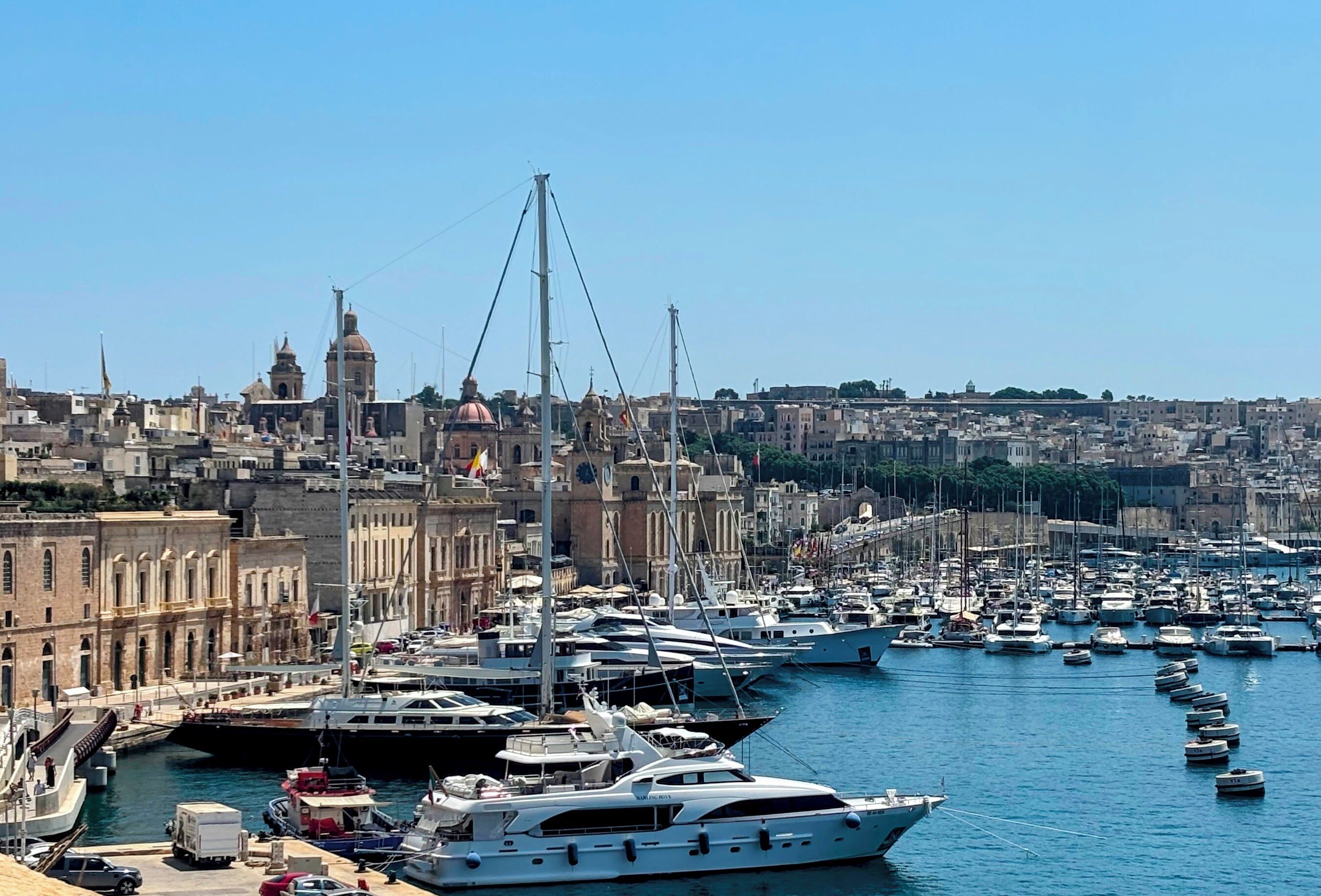 A marina with multiple yachts docked, a prominent church with a dome, and a densely packed urban backdrop.