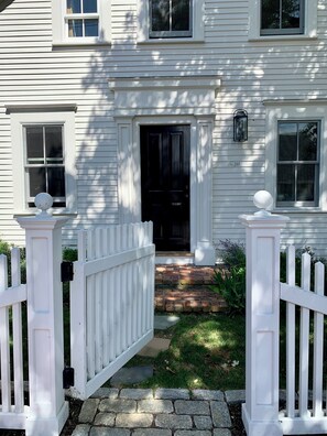 A traditional two-story house with a central door, flanked by white picket fences, features two windows with lanterns above.