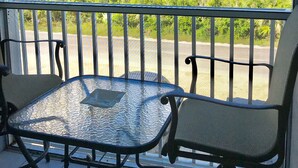 Ein Balkon mit Glastisch und zwei Stühlen, der Blick auf einen Strand mit Vegetation und einem klaren Himmel bietet.