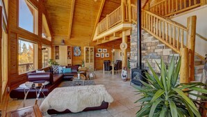A spacious log cabin interior with a stone fireplace, a wooden staircase, and a large window overlooking the outdoors. - Huge Mountain Ski & Hike - Wildernest Haven (Silverthorne)