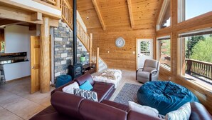 A spacious living area with a wooden ceiling, a stone fireplace, a sofa, a chair, and a large window offering a view of the outdoors. - Huge Mountain Ski & Hike - Wildernest Haven (Silverthorne)