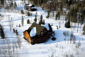 Lumihunnun peittämä hirsimökki, jonka ympärillä on metsää ja taivaalla ei ole pilviä.