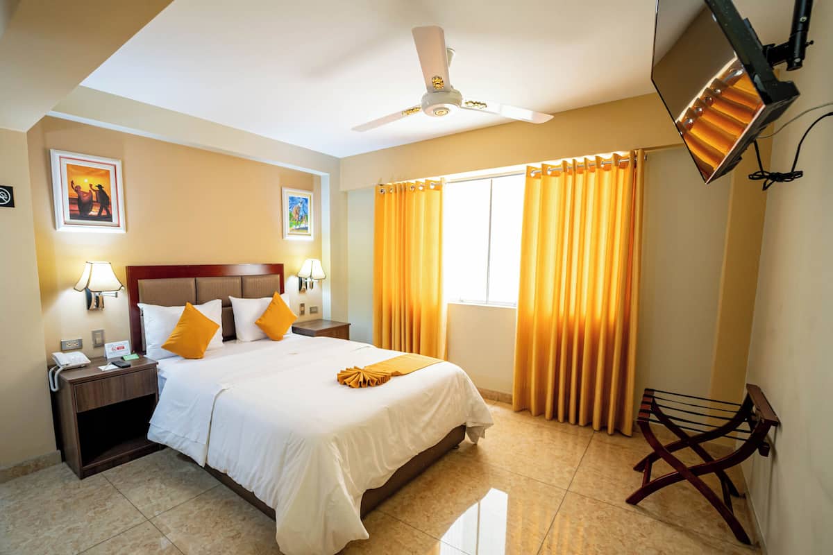 A hotel room with a bed, nightstands, a desk, a chair, a ceiling fan, and a TV mounted on the wall.