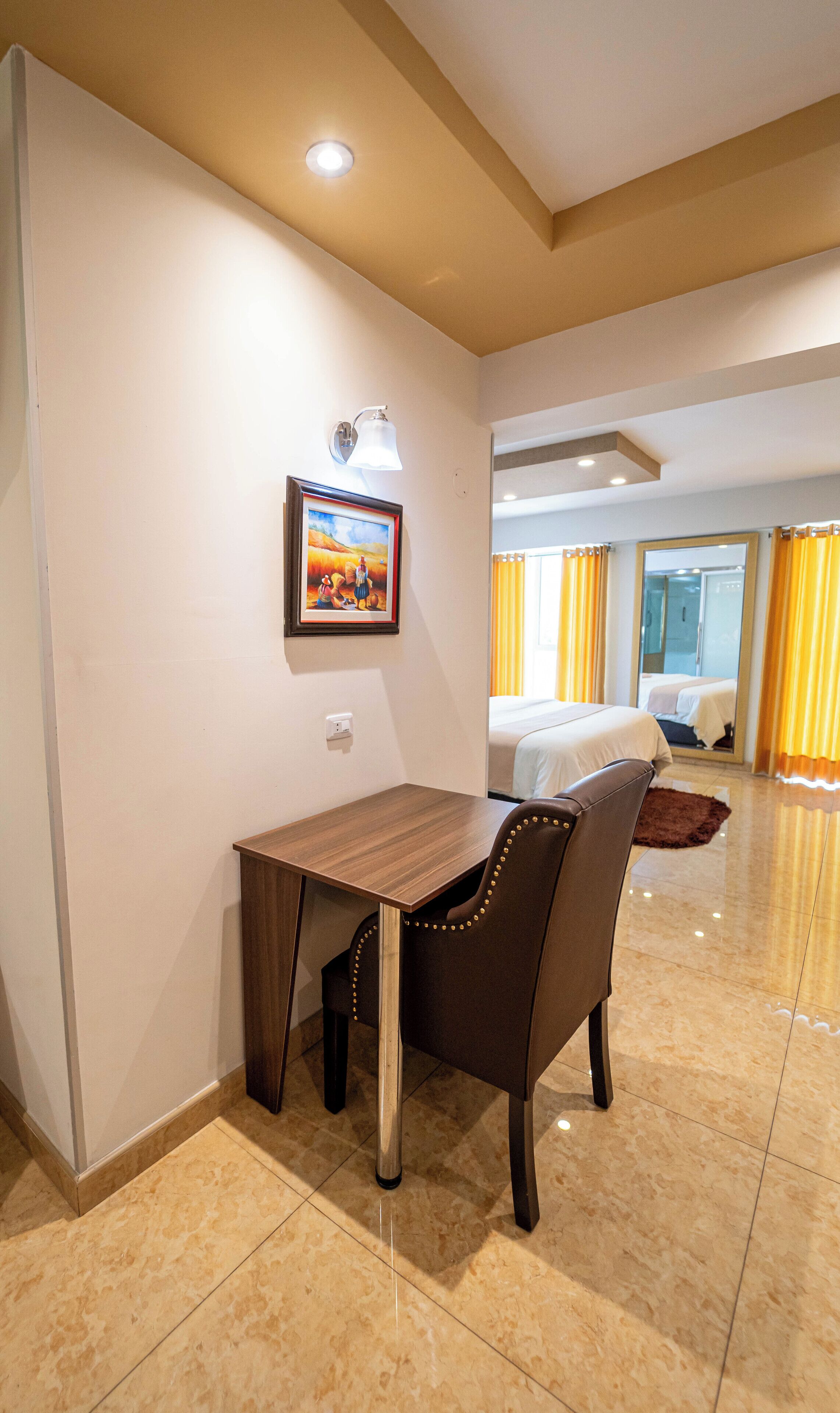 Una habitación de hotel con un escritorio de madera, una silla con tachuelas decorativas, un cuadro enmarcado en la pared y una cama con cortinas amarillas.