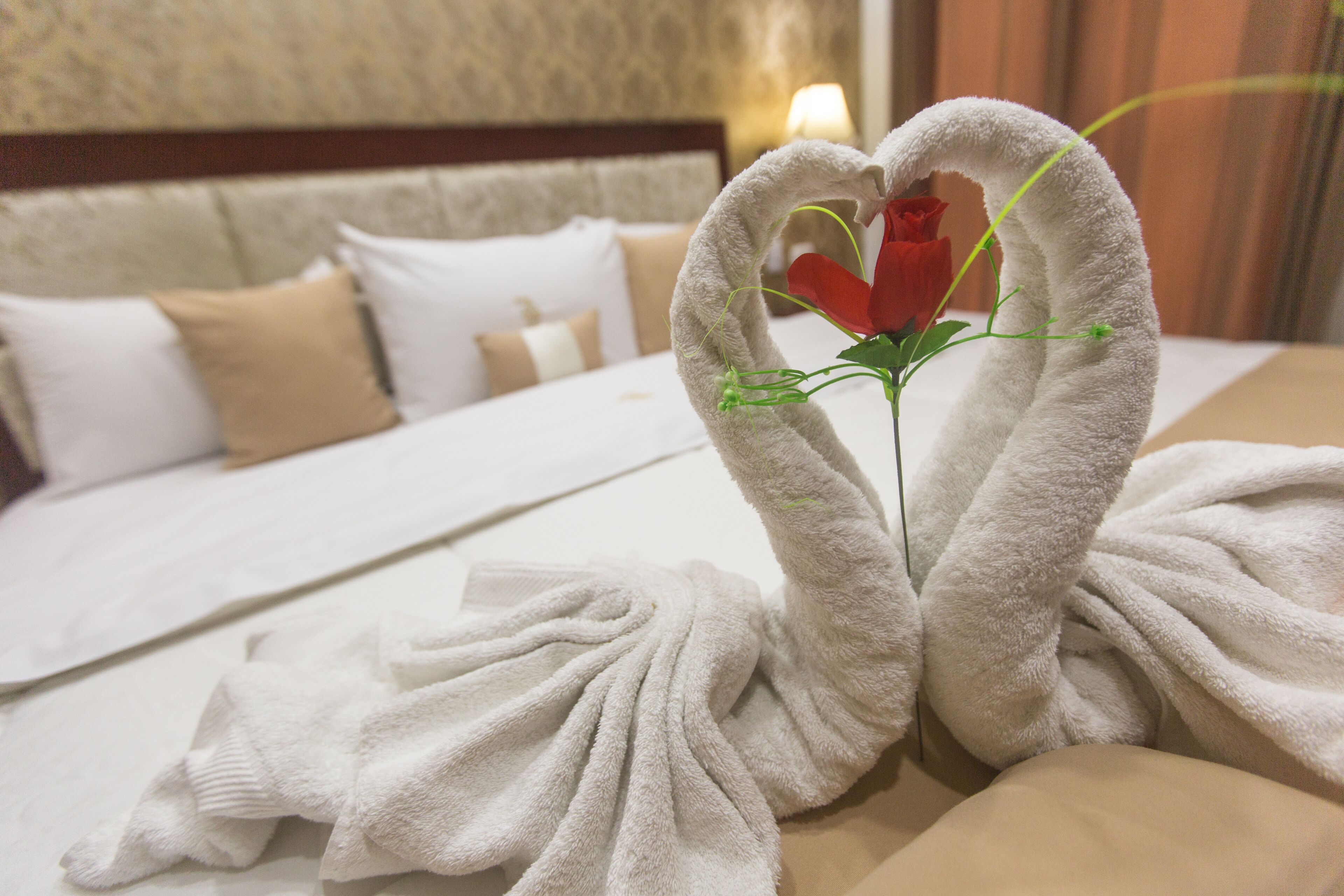 A neatly made bed with white linens and a heart-shaped towel arrangement with a rose.