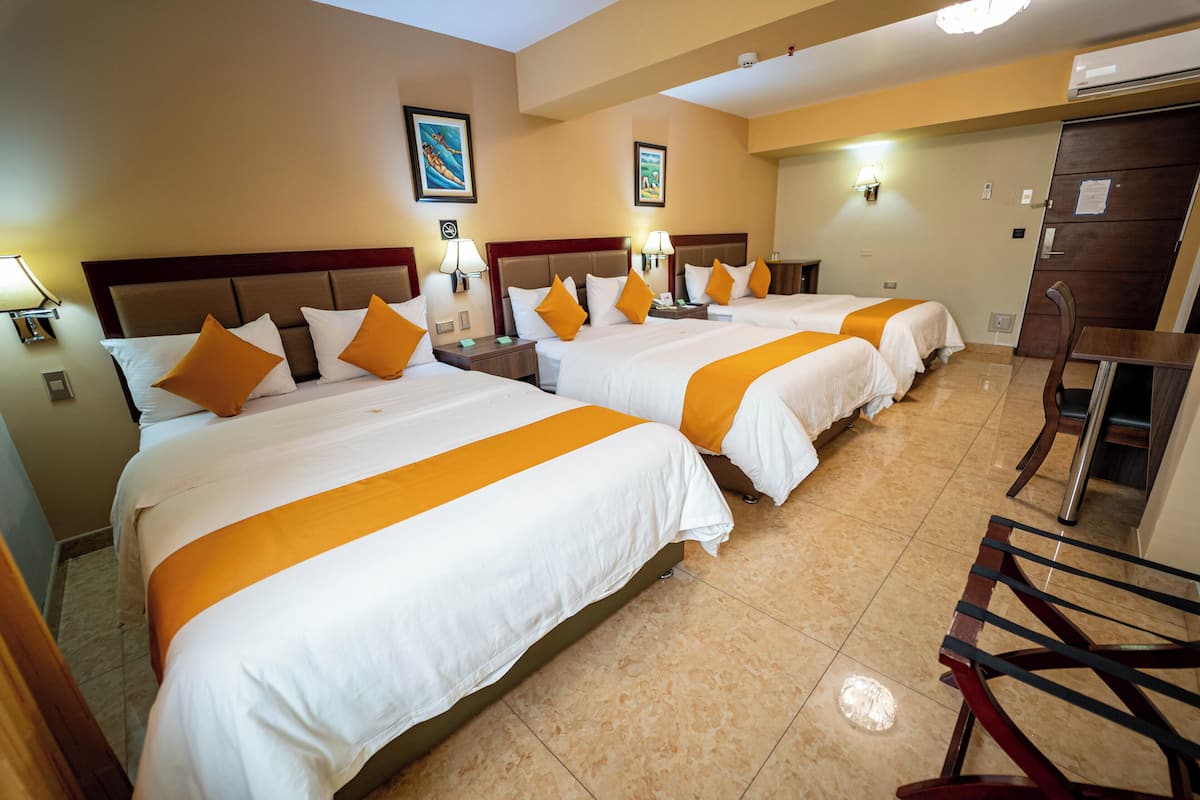 A hotel room with three beds, each with white bedding and orange accents, a wooden headboard, and a small desk with a chair.