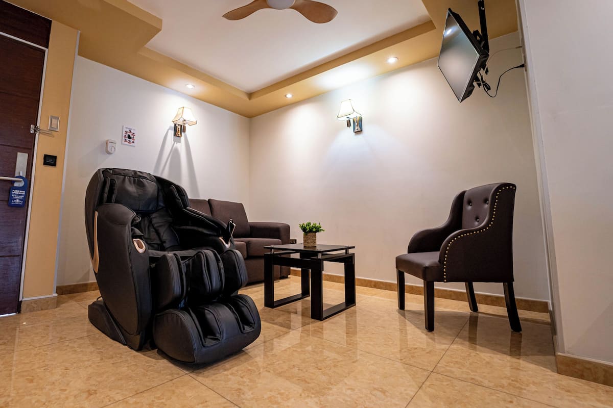 A massage chair, a sofa, a small table with a plant, and a TV mounted on the wall in a room with beige walls and a ceiling fan.