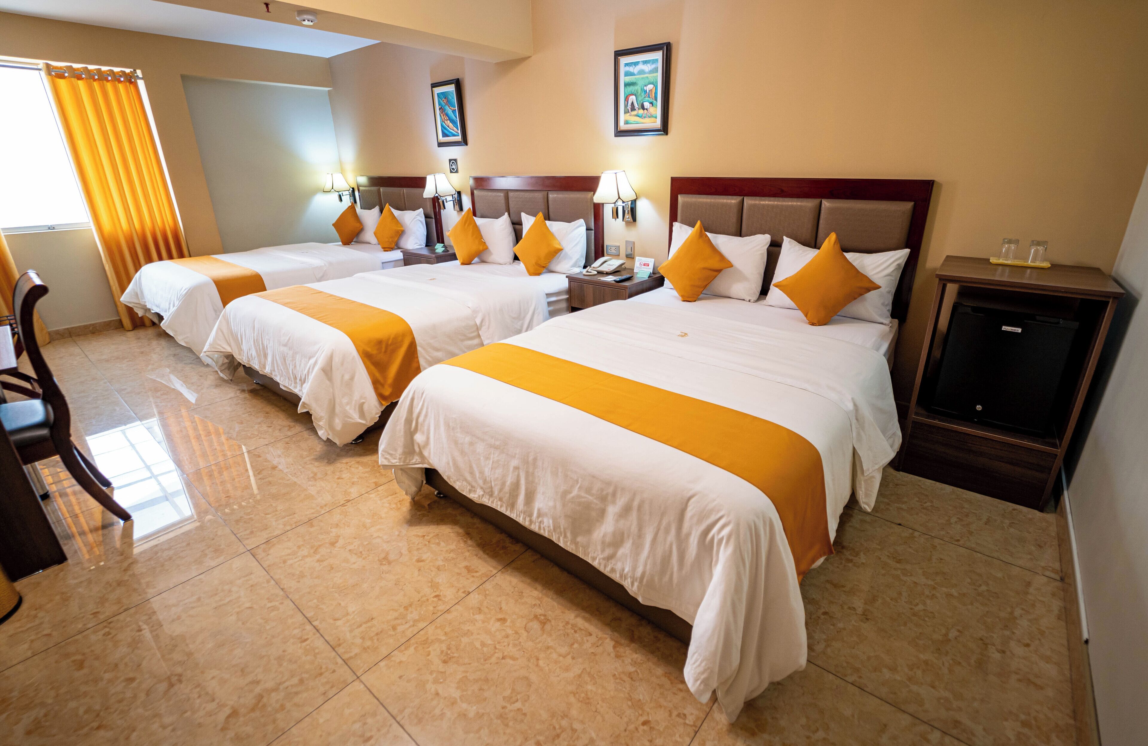 Une chambre d’hôtel avec quatre lits, chacun doté de draps blancs et de touches de couleur orange, d’une tête de lit en bois, d’un bureau avec une chaise et d’une petite armoire.