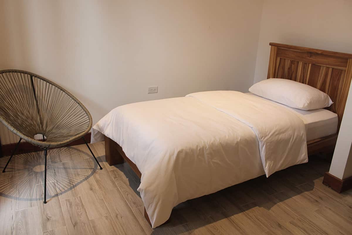 Une chambre avec un lit simple, une tête de lit en bois, une chaise en osier et un plancher en bois.