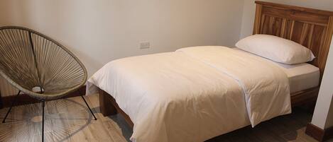 A bedroom with a single bed, a wooden headboard, a woven chair, and a wooden floor.