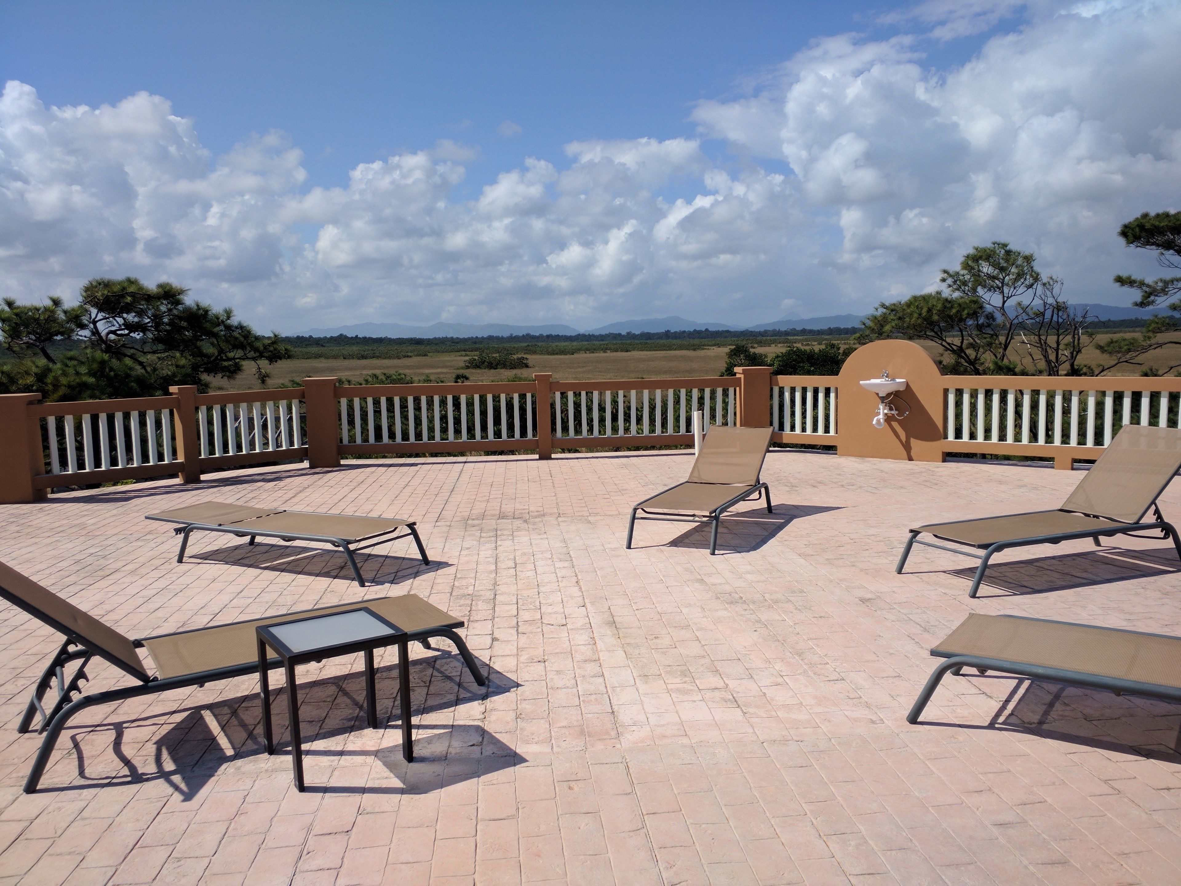 Une terrasse extérieure avec quatre chaises longues et une table, entourée d’une balustrade blanche, offrant une vue panoramique sur un paysage magnifique avec des montagnes et des arbres.