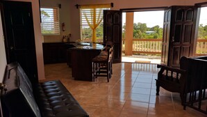 A spacious living area with a dark brown sofa, a dining table with chairs, a kitchen area with a sink and window, and a balcony with a view of greenery. - Savannah Sand Dollar (Hopkins)