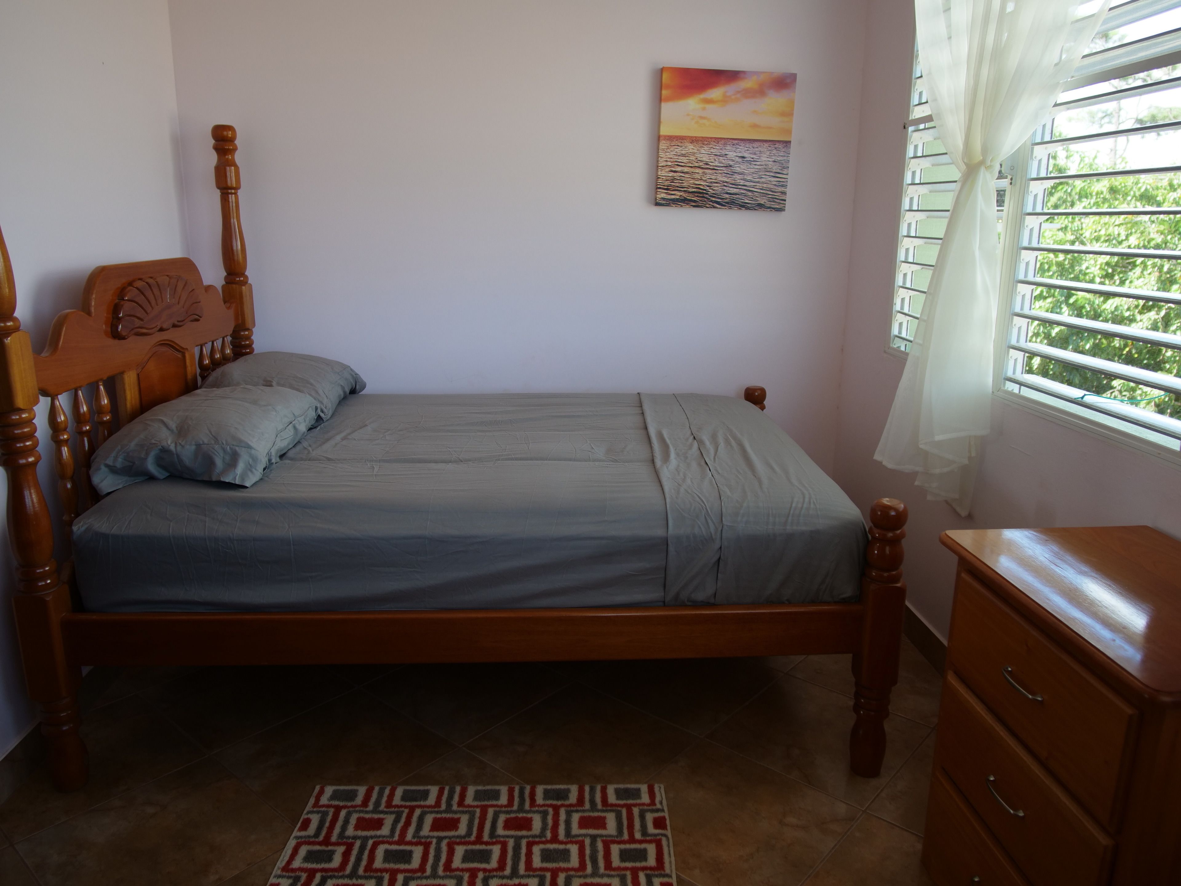A bedroom with a wooden bed, a bedside table, a window with blinds, and a painting on the wall.