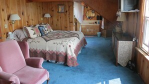 Ein gemütliches Zimmer mit einem Bett, einem rosa Sessel, einer kleinen Küchenzeile und einem Fernseher.