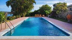 Una piscina rectangular con agua cristalina azul, rodeada por una solana pavimentada. Hay sillones de madera alineados a lo largo del borde de la piscina.