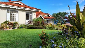 Una casa con techo de tejas rojas, contraventanas blancas y un césped bien cuidado con diversas plantas y flores.