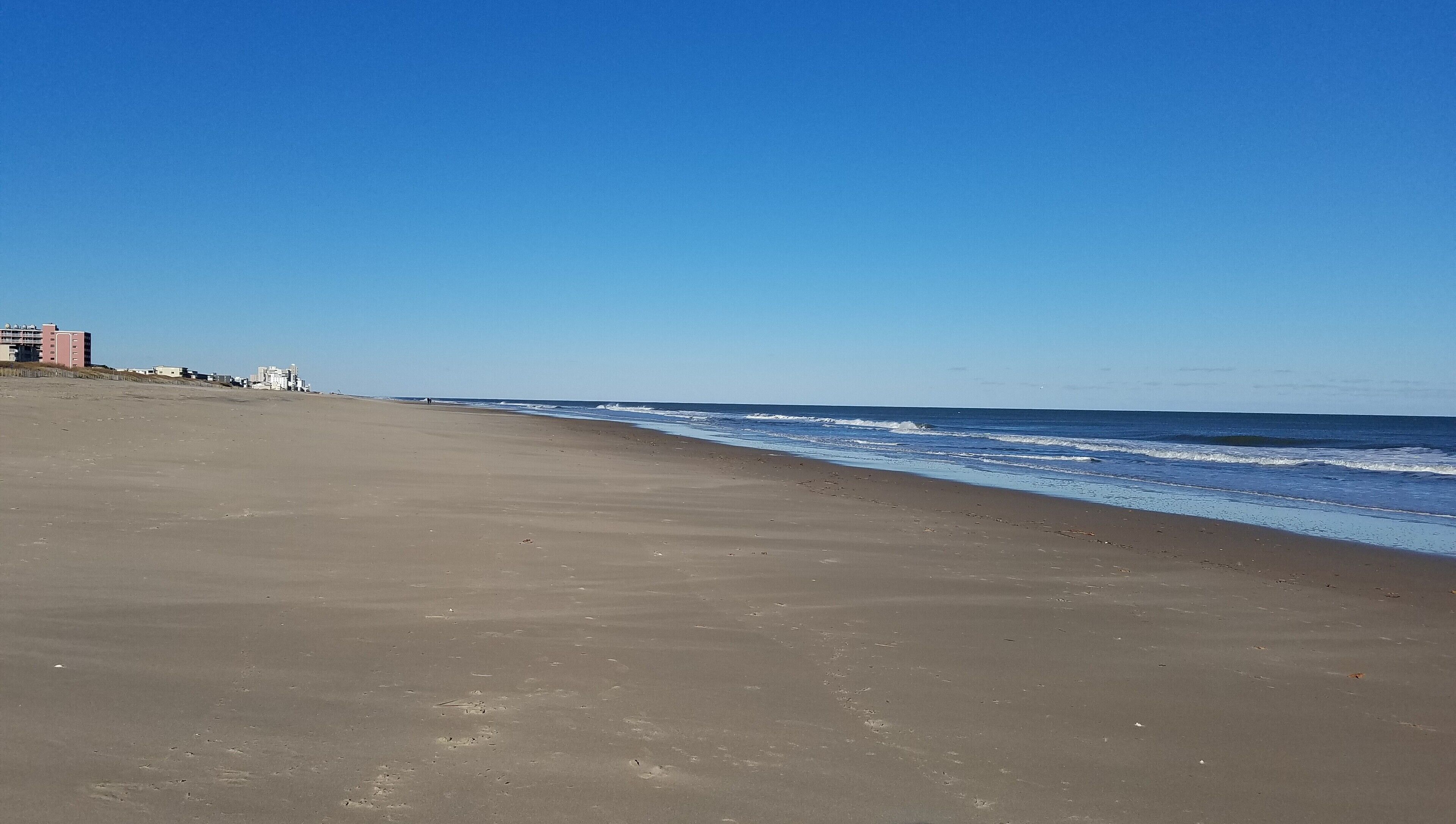 Ein weiter Sandstrand mit sanften Wellen, blauem Himmel und Gebäuden im Hintergrund.