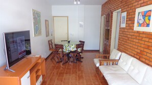 A living room with a TV, a dining table with chairs, a white sofa, and a brick wall with artwork. - Apartment with a balcony just one block from Forte beach! (Cabo Frio)