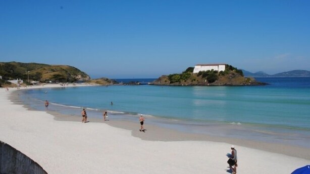 A beach with clear turquoise water, a white sandy shore, and a white building on a small island. - Apartment with a balcony just one block from Forte beach! (Cabo Frio)