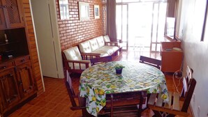 A dining area with a round table covered by a floral tablecloth, surrounded by wooden chairs. There is a brick wall with framed pictures above the sofa. - Apartment with a balcony just one block from Forte beach! (Cabo Frio)