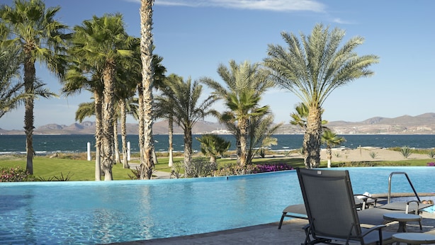 A swimming pool with lounge chairs and palm trees, overlooking a beach and mountains. - BeachFront, Golf, Heated pool, Tennis,Netflix, Gym, Pickleball (La Paz)