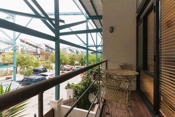 Un balcon doté d’une rambarde en métal, d’une table en osier et offrant une vue sur une rue animée, avec des voitures et des immeubles.
