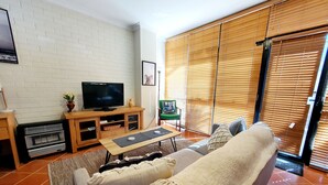 A living room with a sofa, TV, wooden entertainment unit, and a sliding glass door with blinds. - Charming Townhouse in the Heart of Fremantle (Fremantle)
