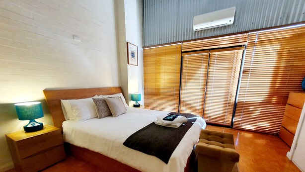 A bedroom with a wooden bed, a nightstand with a lamp, a small sofa, and a window with blinds. - Charming Townhouse in the Heart of Fremantle (Fremantle)