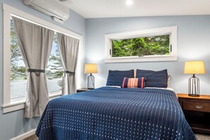 A bedroom with a large bed, two bedside lamps, a window with a view of trees, and an air conditioning unit.