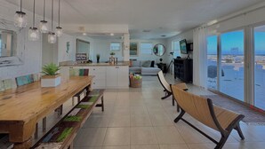 A modern kitchen with a wooden bench, a dining table, and a view of the beach through large sliding doors.