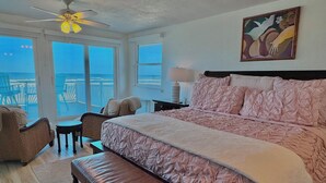 A bedroom with a large bed, a bench, a chair, a table, and a ceiling fan. There is a painting on the wall and a view of the ocean through the windows.