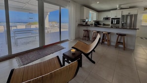 A modern kitchen with white cabinets, a stainless steel refrigerator, and a microwave. There is a dining area with a bench and stools, and a large sliding glass door leading to a balcony with a view of the ocean.