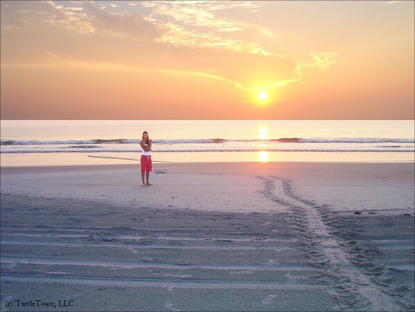 Une scène de plage au coucher du soleil, avec une personne debout sur le sable et des empreintes de pas qui mènent vers l’eau.